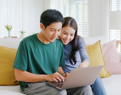 Young couple smiling while booking a hotel on a laptop at home.
