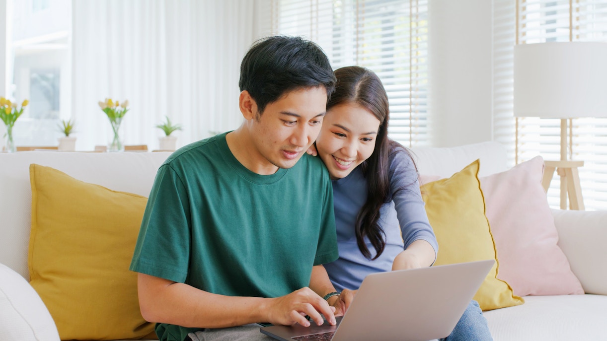 Young couple smiling while booking a hotel on a laptop at home.
