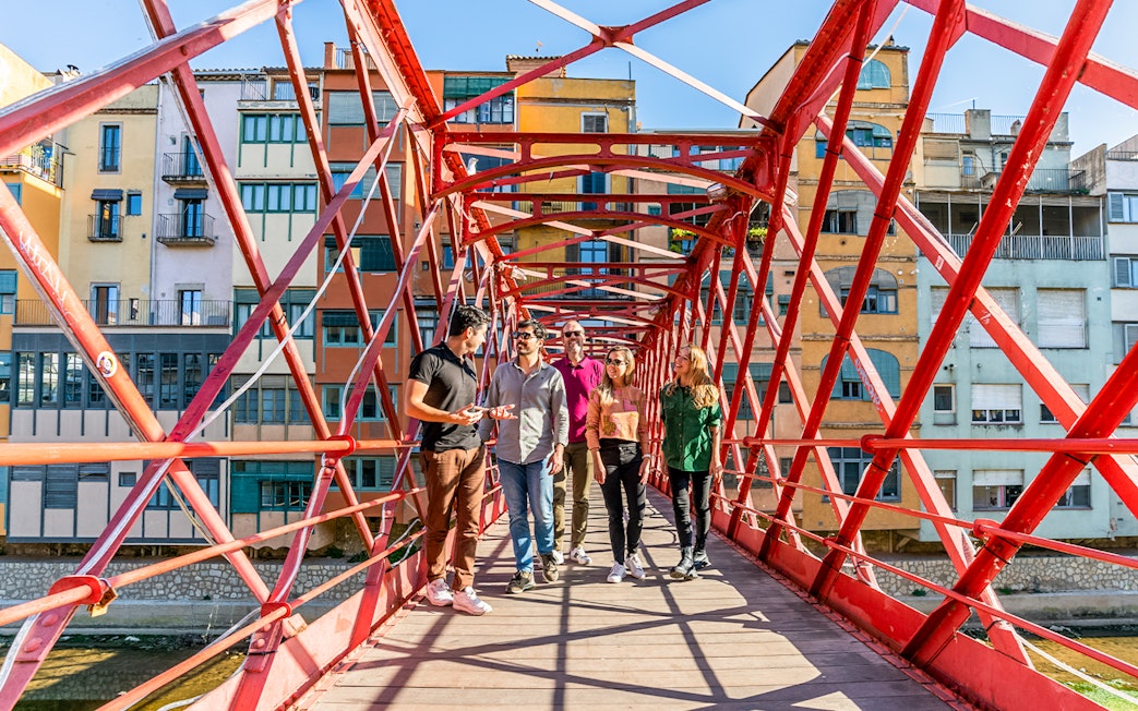 Tourists with guide on Eiffel Bridge in Girona, colorful buildings in background.