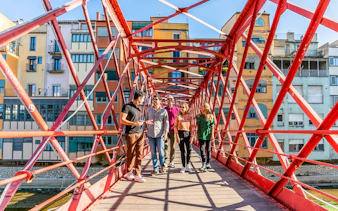 Tourists with guide on Eiffel Bridge in Girona, colorful buildings in background.