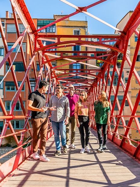 Tourists with guide on Eiffel Bridge in Girona, colorful buildings in background.