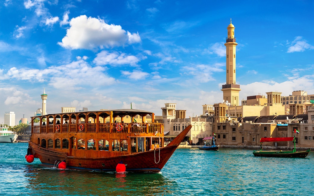 Traditional dhow boat cruising on Dubai Creek with cityscape and minaret in the background.