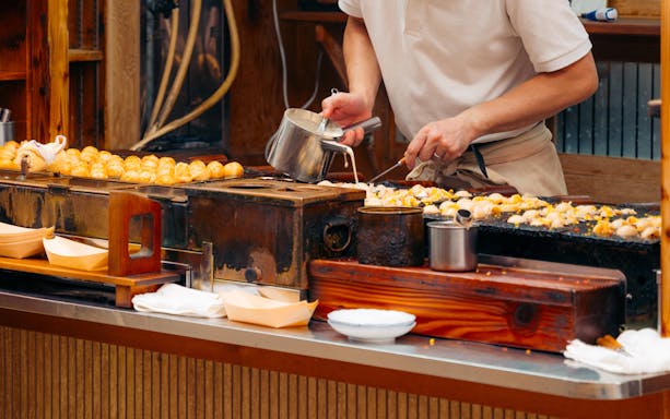 Osaka chef cooking takoyaki on a grill at a street food stall.