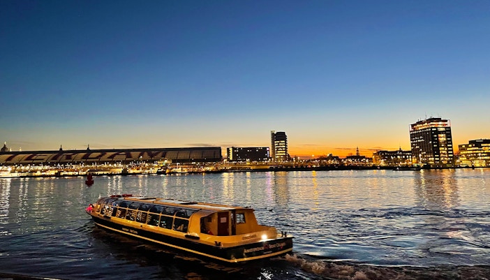 Amsterdam canal cruise boat at sunset with city skyline in the background.