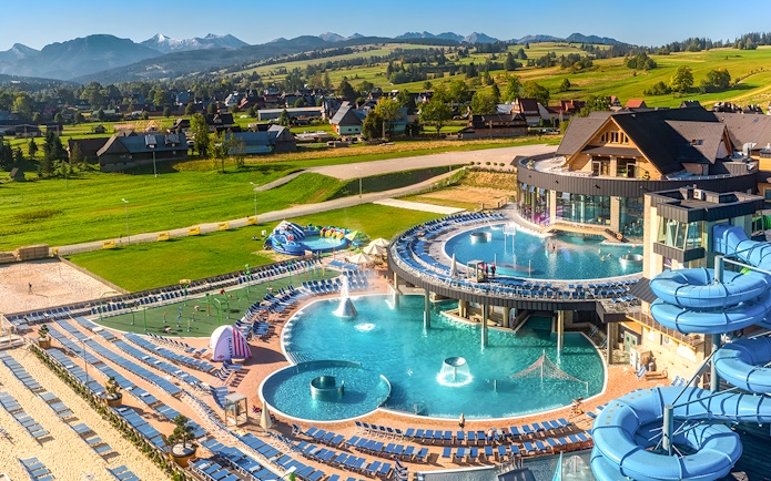 Tourists relaxing in a thermal spa with pools and slides in Zakopane, Poland, surrounded by scenic mountains.