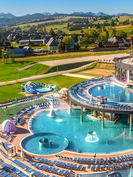 Tourists relaxing in a thermal spa with pools and slides in Zakopane, Poland, surrounded by scenic mountains.