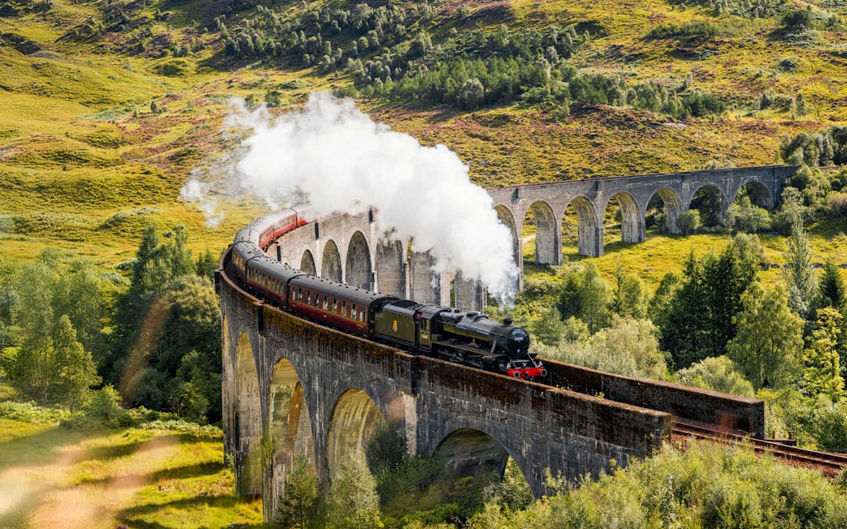 Steam train crossing Glenfinnan Viaduct in Scotland with lush green hills in the background.