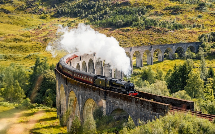 Steam train crossing Glenfinnan Viaduct in Scotland with lush green hills in the background.
