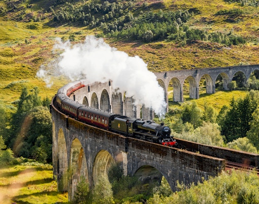 Steam train crossing Glenfinnan Viaduct in Scotland with lush green hills in the background.