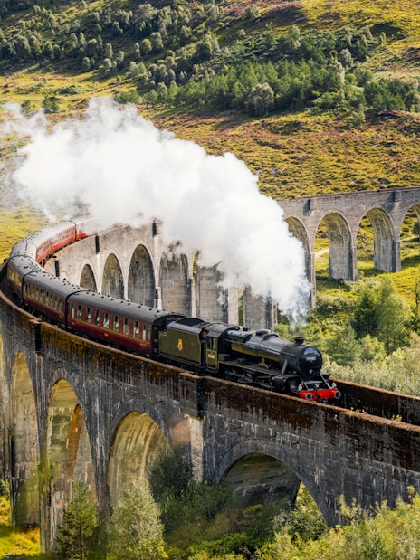 Steam train crossing Glenfinnan Viaduct in Scotland with lush green hills in the background.