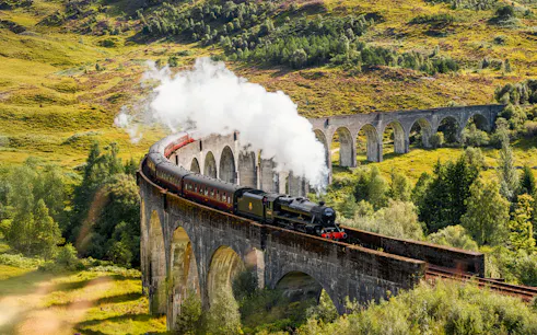 Steam train crossing Glenfinnan Viaduct in Scotland with lush green hills in the background.