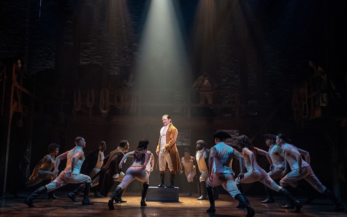 Stage performance of Hamilton with actors in period costumes under spotlight.