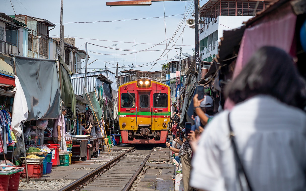 Train passing through Maeklong Railway Market, Bangkok, with vendors and tourists nearby.