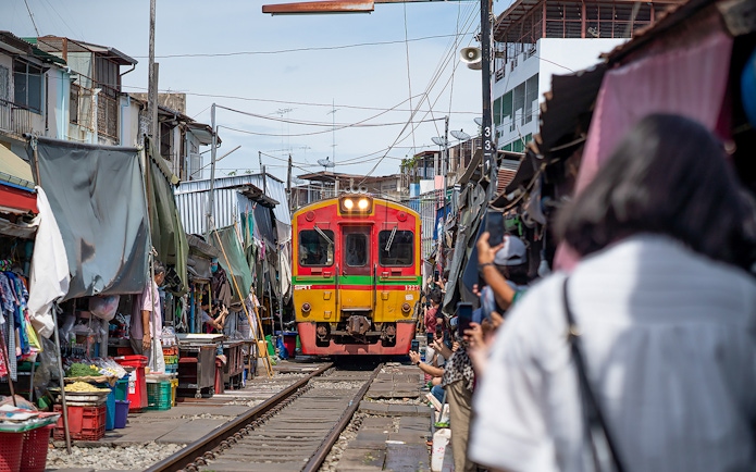 Train passing through Maeklong Railway Market, Bangkok, with vendors and tourists nearby.