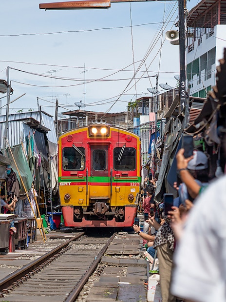 Train passing through Maeklong Railway Market, Bangkok, with vendors and tourists nearby.