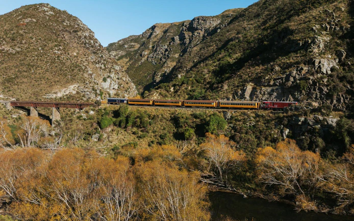 Taieri Gorge train crossing Deep Stream Bridge with rocky hills and autumn trees in New Zealand.