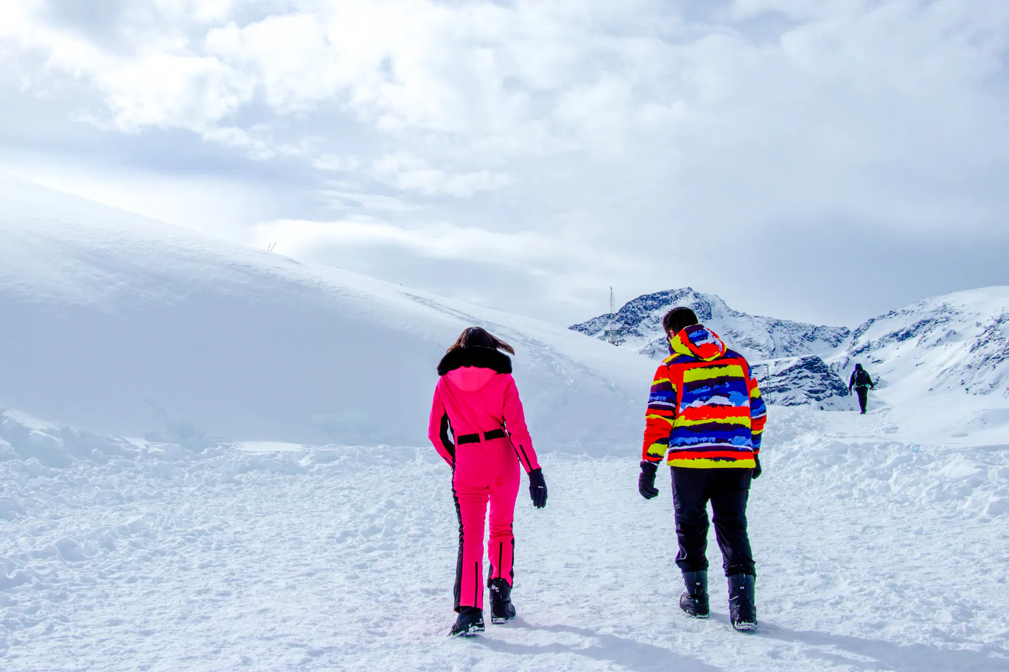 Two people walking on a snowy path in Valle Nevado, Santiago, with mountains in the background.