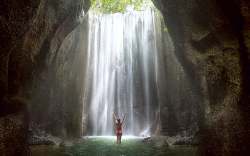 Woman standing in front of a waterfall in Bali, arms raised, surrounded by lush cliffs.