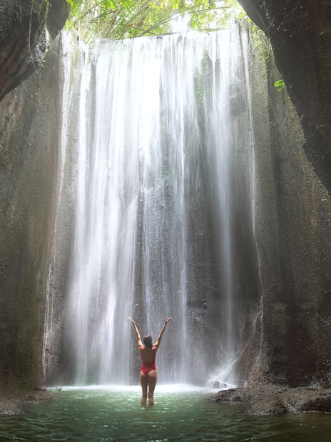 Woman standing in front of a waterfall in Bali, arms raised, surrounded by lush cliffs.