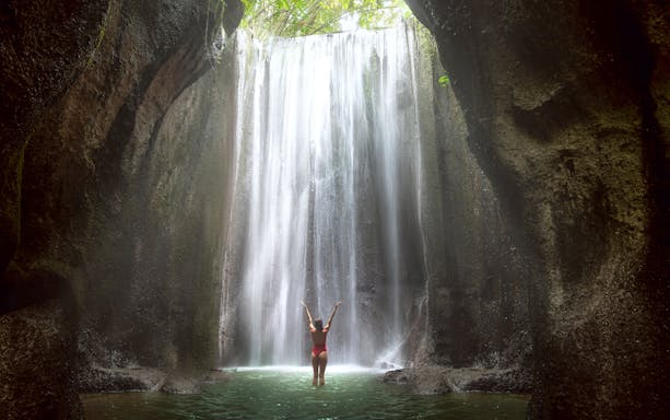 Woman standing in front of a waterfall in Bali, arms raised, surrounded by lush cliffs.