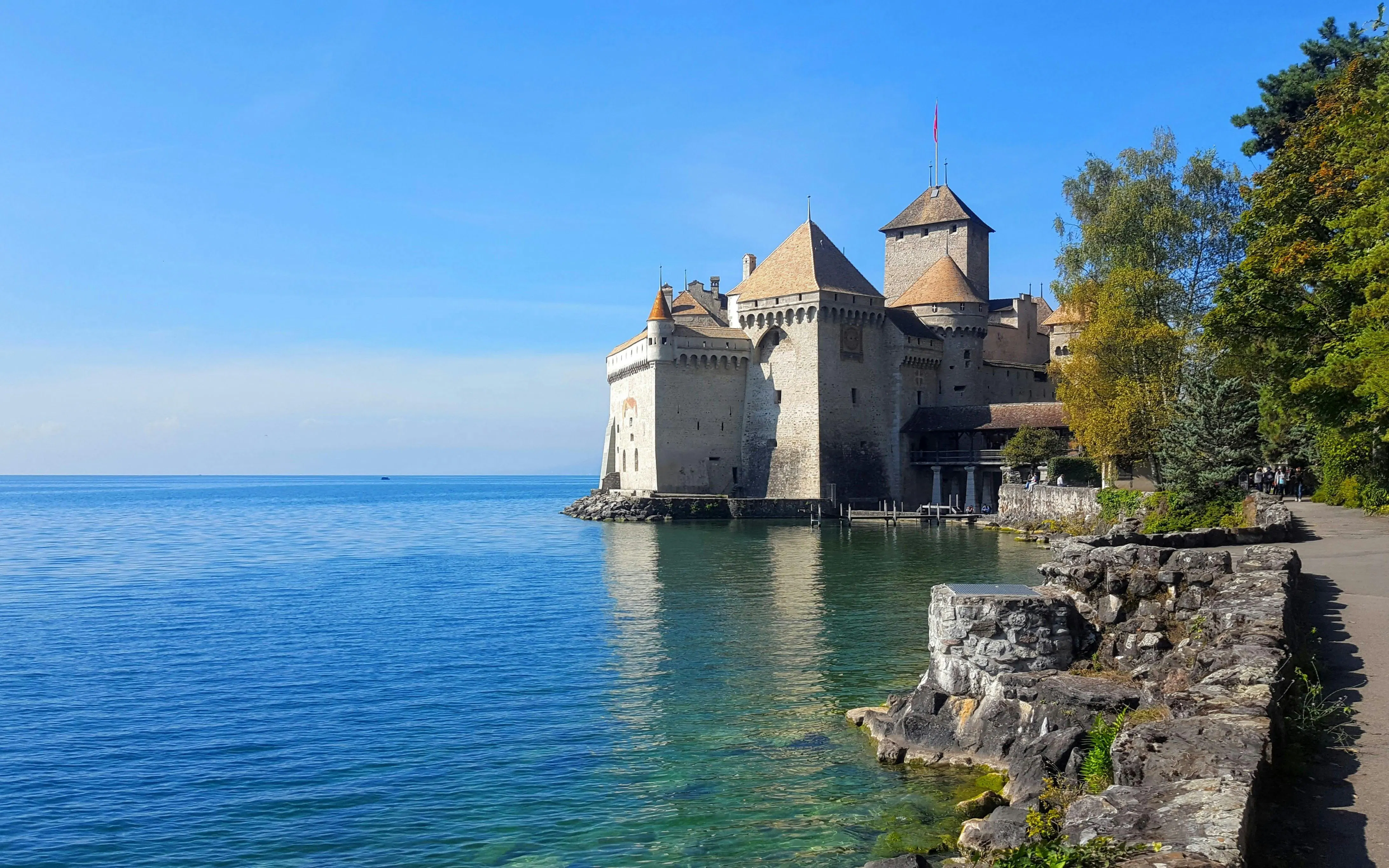 Chillon Castle on Lake Geneva with surrounding trees and clear blue water.