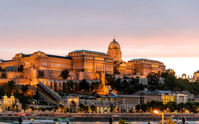 Buda Castle illuminated at night in Budapest, Hungary, with a sunset sky.