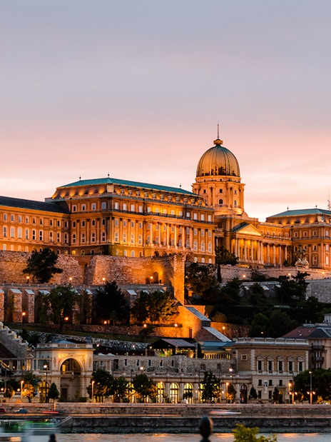 Buda Castle illuminated at night in Budapest, Hungary, with a sunset sky.