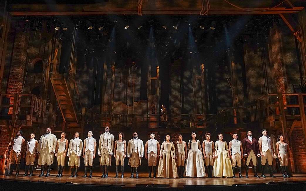 Cast of Hamilton performing on stage with period costumes and set design.