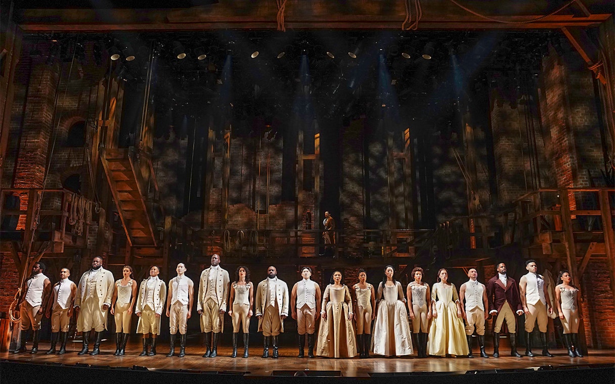 Cast of Hamilton performing on stage with period costumes and set design.