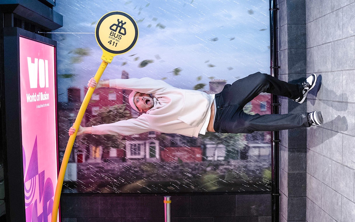 Guest clinging to a tilted bus stop sign at World of Illusions, creating a gravity-defying effect.