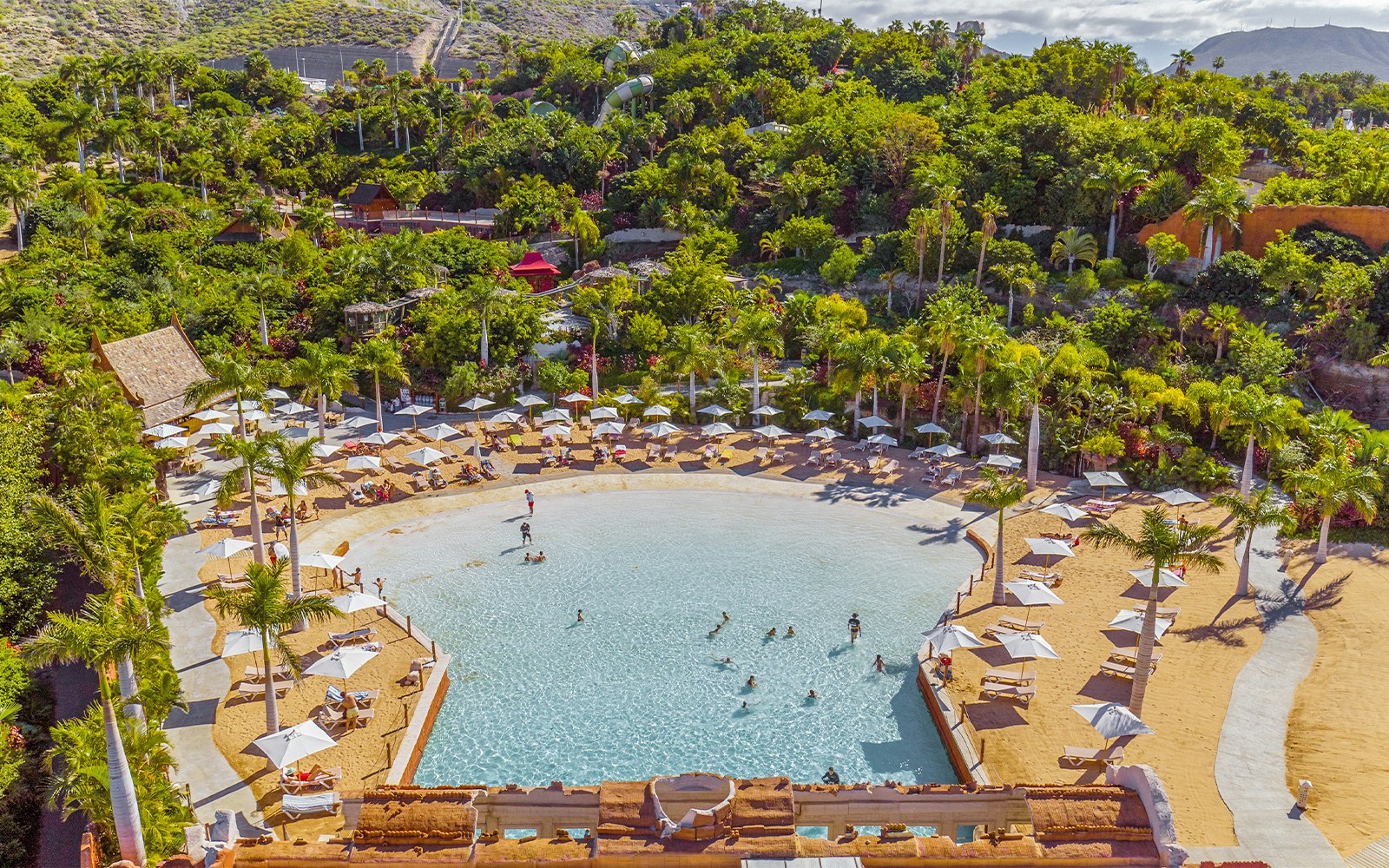 Tourists relaxing on Coco Beach at Siam Park with palm trees and water slides in the background.