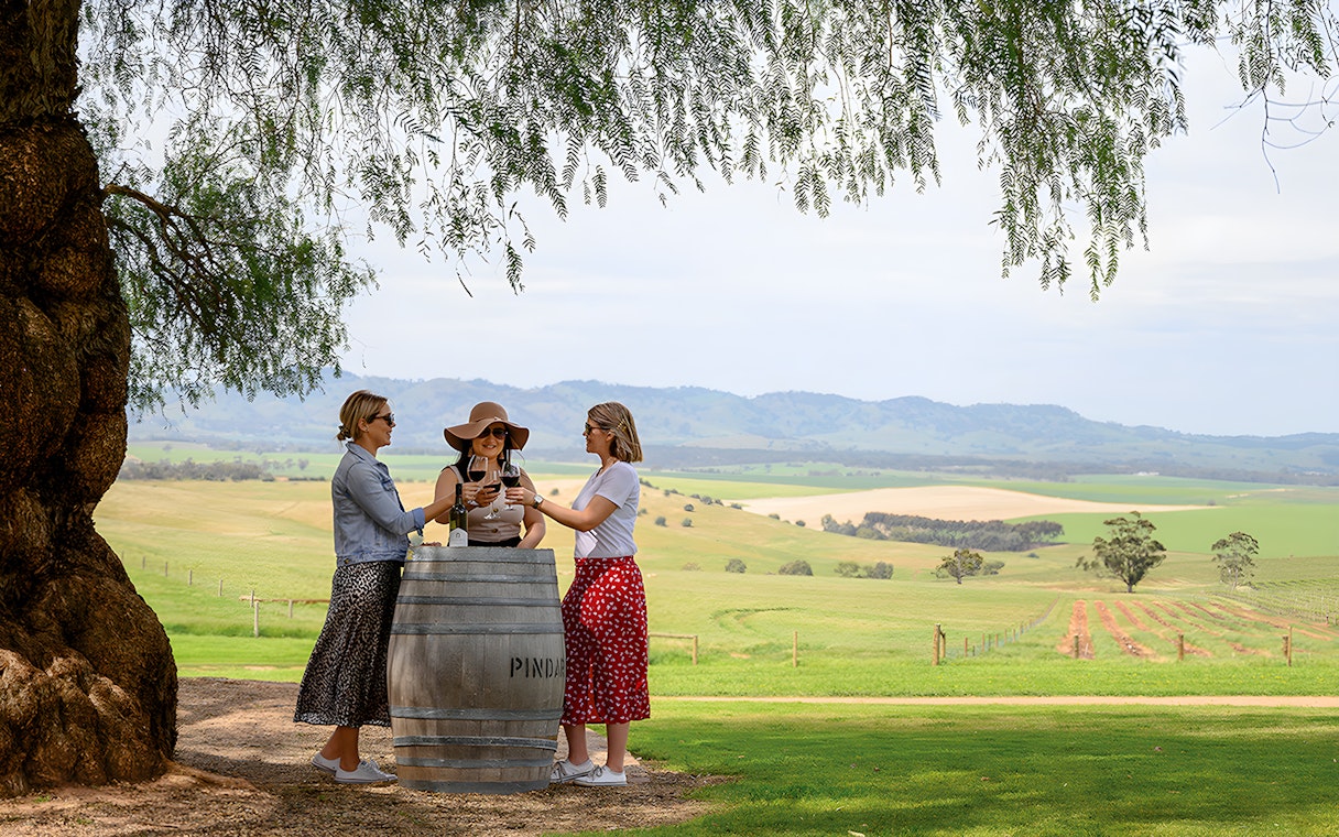 Three people enjoying wine at a barrel table with Barossa Valley vineyards in the background.