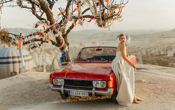 Classic car with a woman in a dress, Cappadocia landscape and hot air balloons in the background.