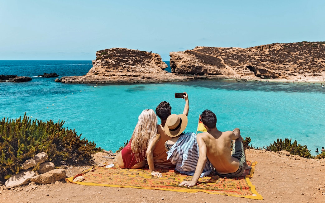 Friends taking a selfie at Blue Lagoon, Malta, with clear turquoise water in the background.
