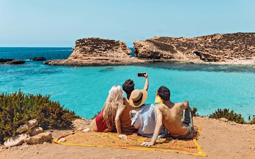 Friends taking a selfie at Blue Lagoon, Malta, with clear turquoise water in the background.