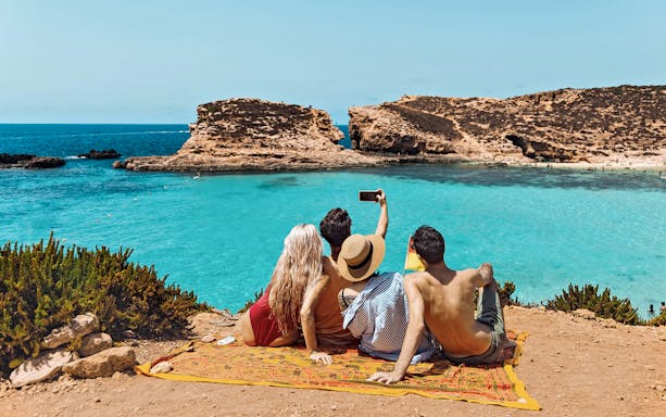 Friends taking a selfie at Blue Lagoon, Malta, with clear turquoise water in the background.