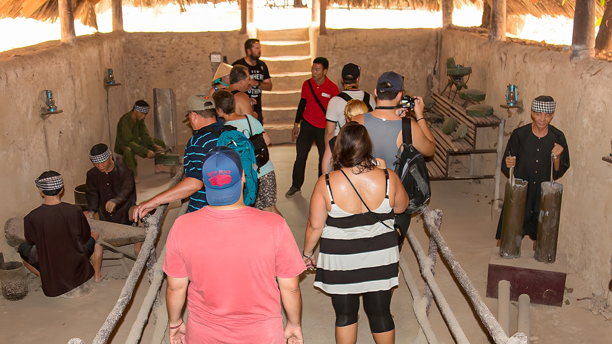 Tour group with guide in Cu Chi tunnel kitchen, observing traditional cooking methods.