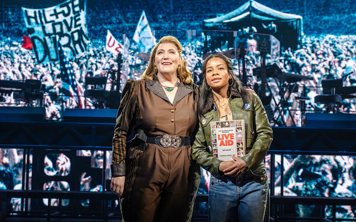 Two performers on stage at Just for One Day, holding a Live Aid book with a concert backdrop.