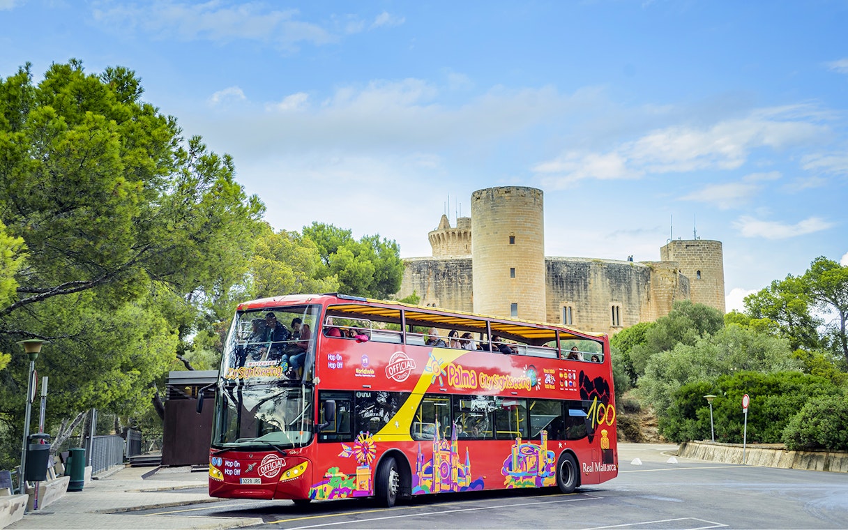 HOHO tour bus in front of Bellver Castle, Mallorca, surrounded by trees.