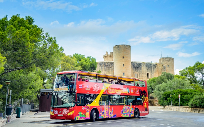 HOHO tour bus in front of Bellver Castle, Mallorca, surrounded by trees.