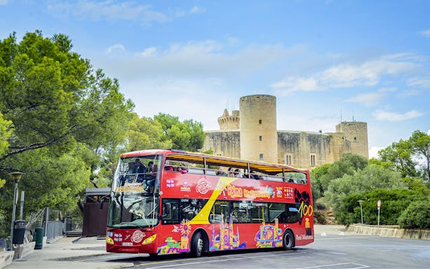 HOHO tour bus in front of Bellver Castle, Mallorca, surrounded by trees.