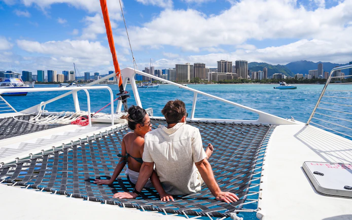 Couple relaxing on a catamaran net with Waikiki skyline in the background during whale watch sail.