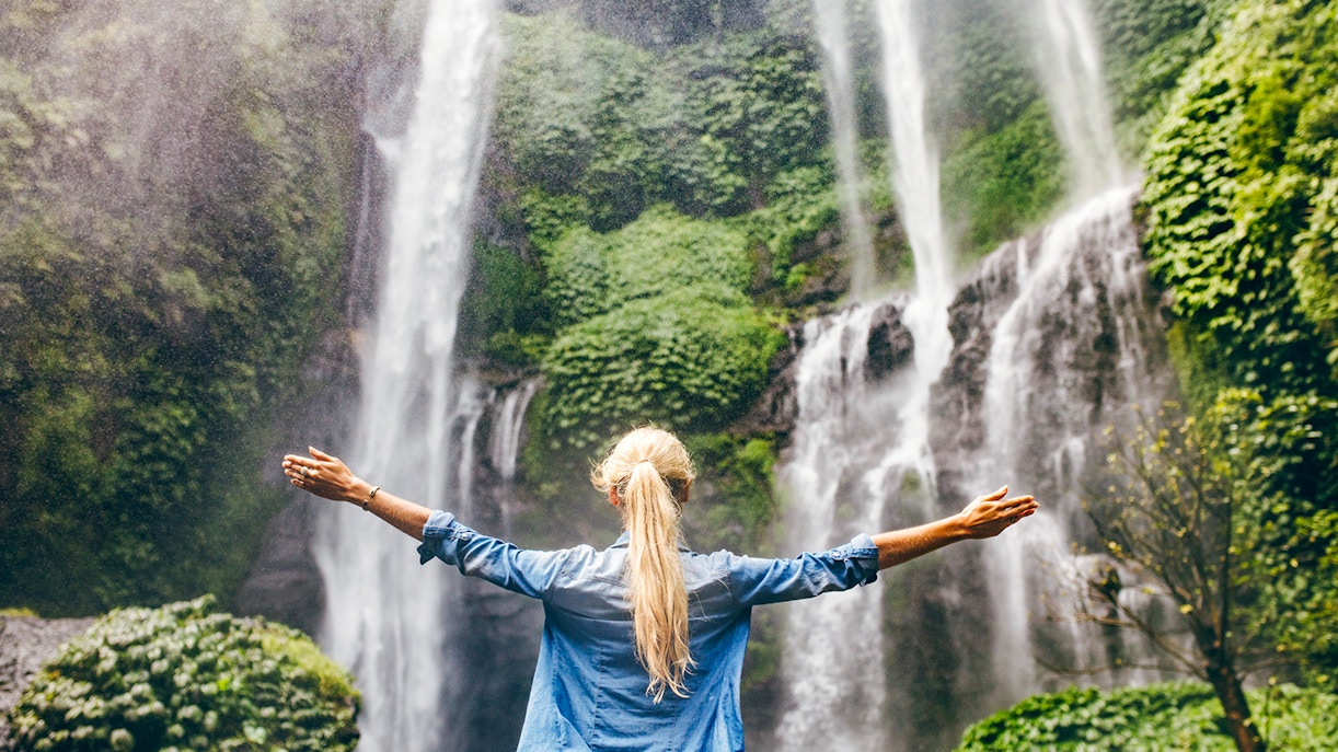 Person standing with arms outstretched in front of a waterfall on the Road to Hana, Maui, Hawaii.
