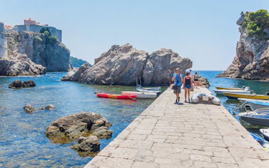 Tourists walking along a stone pier in Dubrovnik with boats and rocky cliffs nearby.
