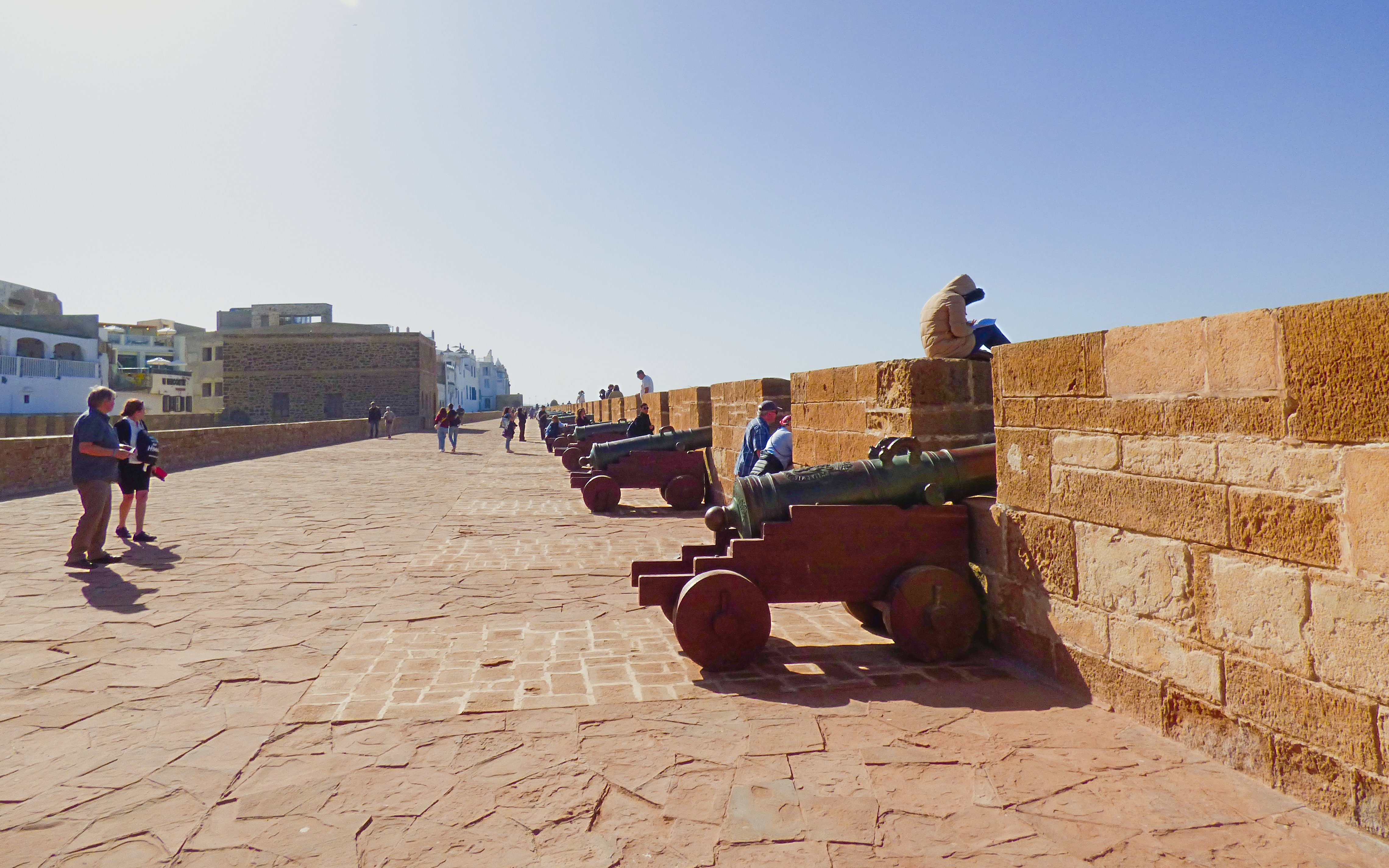 Cannons line the ramparts of Essaouira, Morocco, with people walking and sitting nearby.