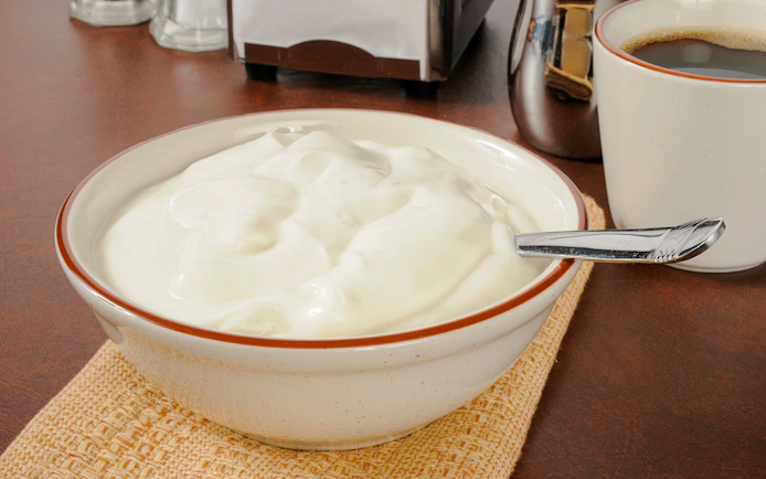 Bowl of plain yogurt with a spoon on a table next to a cup of coffee.
