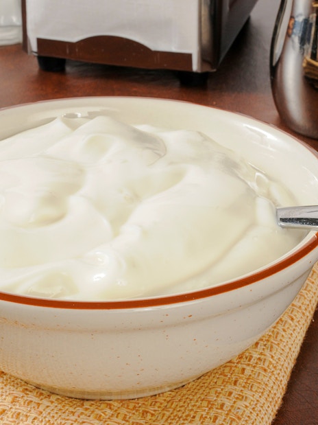 Bowl of plain yogurt with a spoon on a table next to a cup of coffee.
