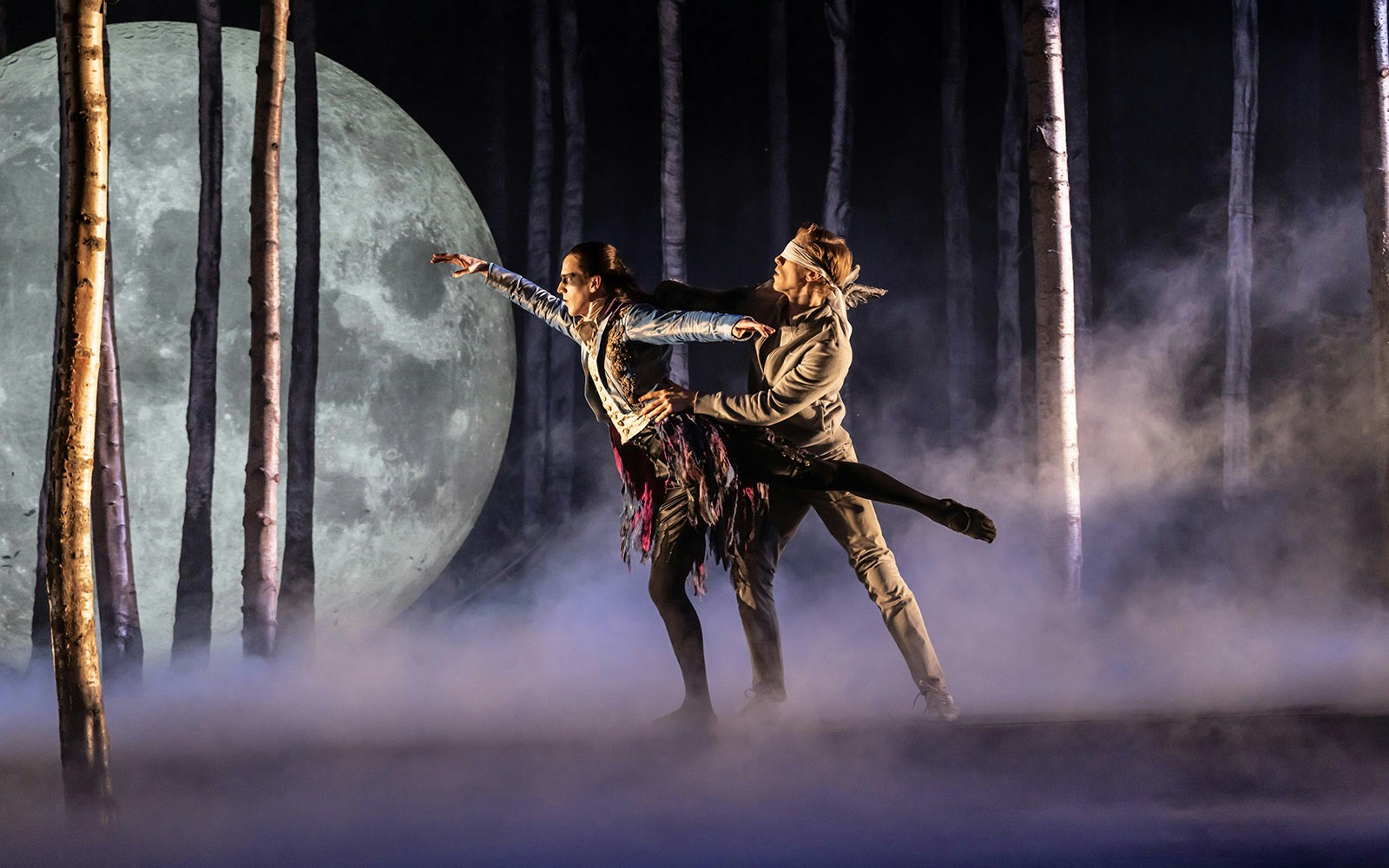 Dancers performing in a forest scene from Matthew Bourne's Sleeping Beauty with a large moon backdrop.