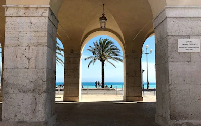 Arched walkway with palm tree and ocean view in Nice, France, part of self-guided city exploration game.