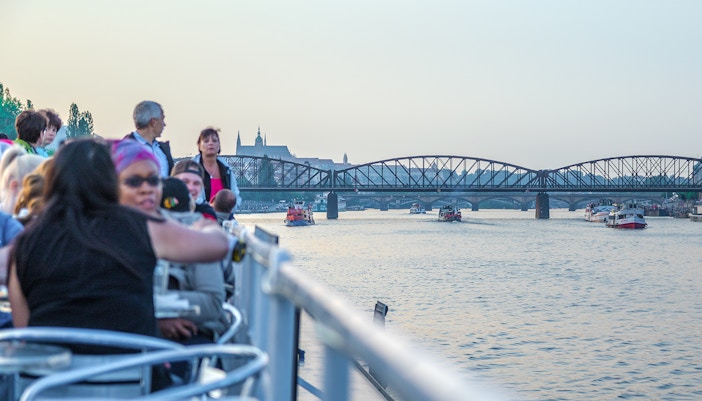 People enjoying a cruise on the Vltava River with a view of a bridge and distant cityscape.